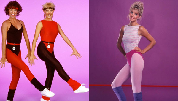 Trois femmes en tenues d’aérobic typiques des années 80 avec justaucorps moulants, leggings contrastés, ceintures fines et jambières colorées sur fond rose, illustrant la mode sportive énergique et flashy de l’époque.